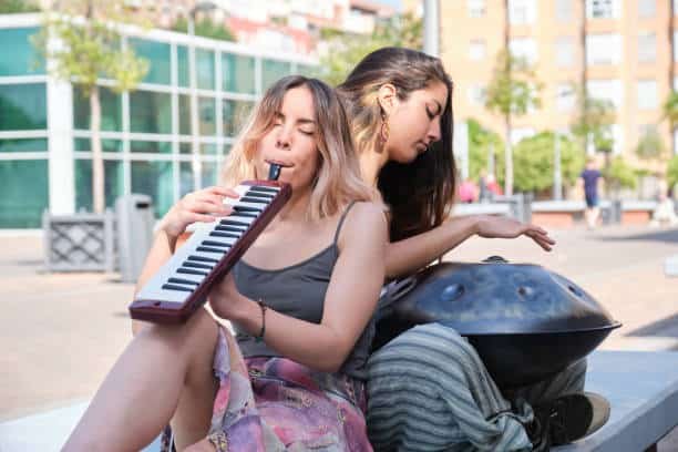 two women playing a handpan and a melodica blow organ sitting at street.